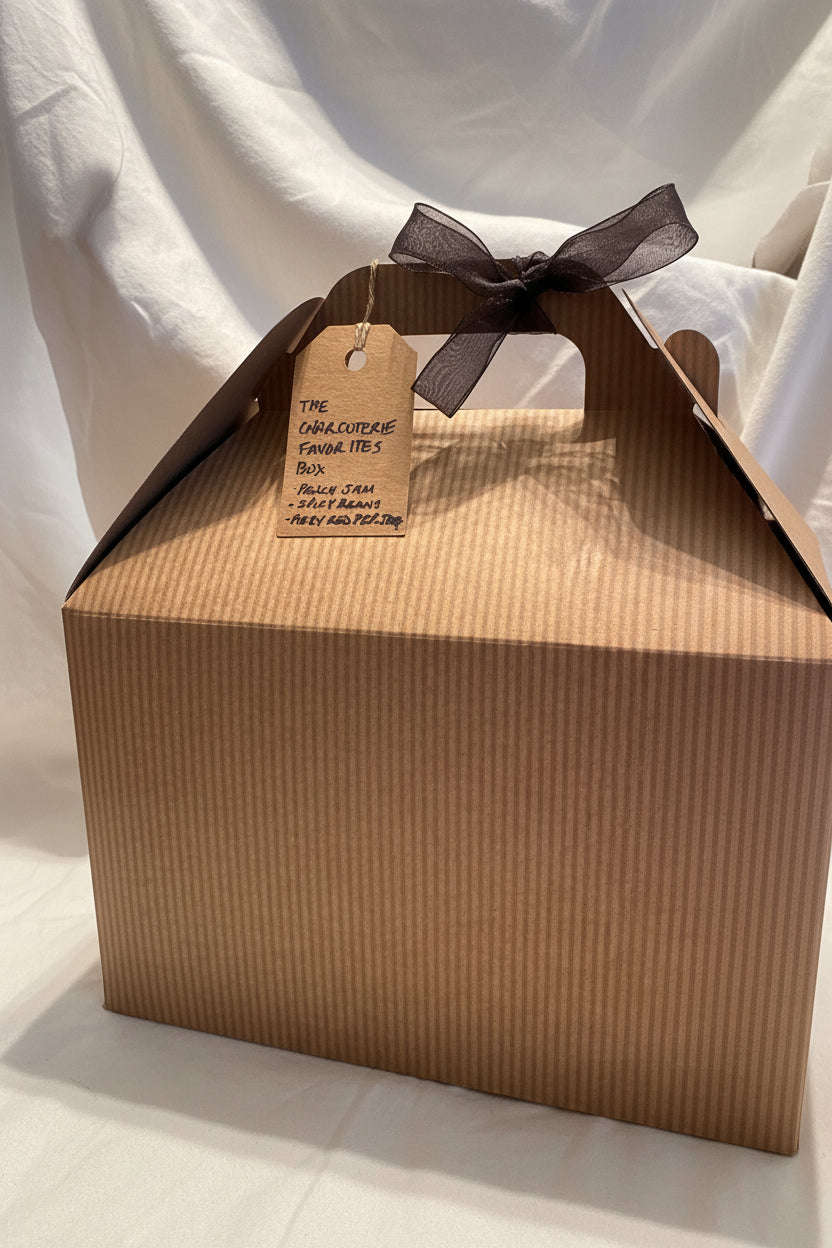 A brown corrugated cardboard gift box with a handle, tied with a dark ribbon, and featuring a kraft paper hangtag that reads 'THE CHAROCUTERIE FAVORITES BOX' with listed items below.